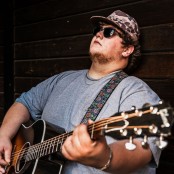 A man leaning against a wooden wall holding a guitar wearing a blue tshirt, camo hat, and black sunglasses